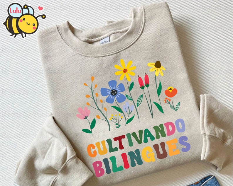 Peut inclure: Un sweat-shirt beige avec un motif floral color&eacute; et le texte "Cultivando bilingues" en lettres arc-en-ciel.
