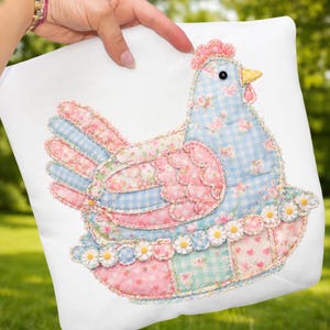 Op de afbeelding: Een wit vierkant kussen met een patchwork kippenontwerp. De kip heeft een blauw geruit lichaam met bloemaccenten, roze vleugels en een roze kam. Het ontwerp is versierd met madeliefjes.