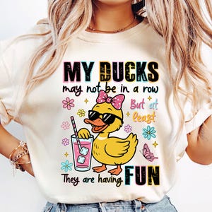 May include: Cream-colored graphic t-shirt with the text "MY DUCKS may not be in a row But at least They are having FUN." The graphic features a yellow duck wearing sunglasses and a pink bow, holding a drink with ice cubes.