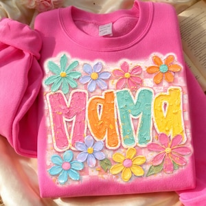 Puede incluir: Una sudadera de cuello redondo en color rosa brillante con la palabra MAMA en letras grandes, texturizadas y multicolores. El diseño incluye un fondo de cuadros rosa claro y varias flores coloridas en tonos azul, morado, rosa, naranja y amarillo.