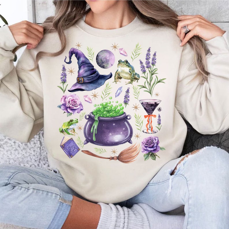 May include: A cream-colored sweatshirt with a whimsical design featuring a witch's hat, a cauldron, frogs, lavender, and a potion. The design is in shades of purple, green, and gold.