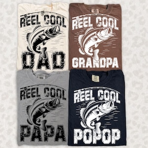 Puede incluir: Cuatro camisetas en crema, marr&oacute;n, gris y azul marino, cada una con un dise&ntilde;o de pesca. El dise&ntilde;o incluye las palabras "Reel Cool" encima de una ilustraci&oacute;n de pez, y las palabras "Dad", "Grandpa", "Papa" y "Popop" debajo.