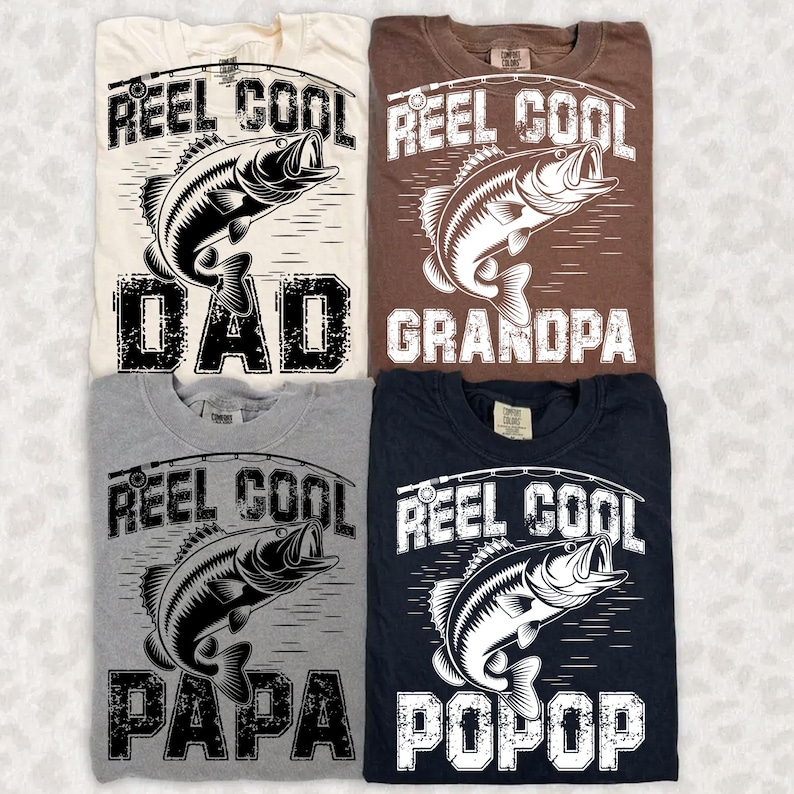 Puede incluir: Cuatro camisetas en crema, marr&oacute;n, gris y azul marino, cada una con un dise&ntilde;o de pesca. El dise&ntilde;o incluye las palabras "Reel Cool" encima de una ilustraci&oacute;n de pez, y las palabras "Dad", "Grandpa", "Papa" y "Popop" debajo.