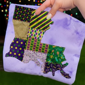 May include: A purple and multi-patterned Louisiana-shaped decorative item. The design incorporates various fabrics with patterns including polka dots, stripes, and fleur-de-lis. The item is held by a hand, with a blurred background of lights.