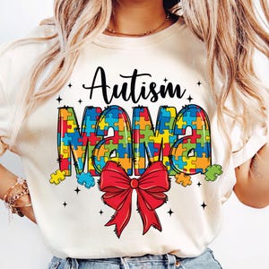 May include: White t-shirt with the text "Autism Mama" in colorful puzzle pieces and a red bow.