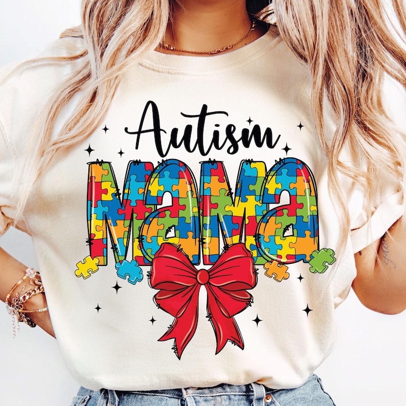 May include: White t-shirt with the text "Autism Mama" in colorful puzzle pieces and a red bow.