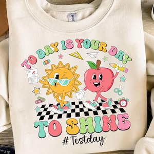 May include: A white sweatshirt with a colorful graphic design featuring a sun and an apple wearing sunglasses and holding hands. The text "TO DAY IS YOUR DAY TO SHINE #Testday" is printed in a retro font.