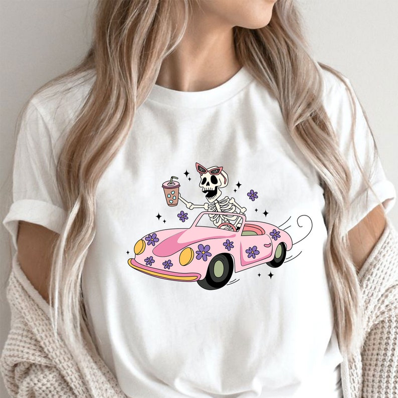 May include: White t-shirt with a graphic of a skeleton wearing sunglasses and driving a pink car with flowers. The skeleton is holding a cup of coffee.
