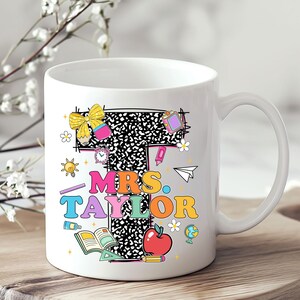 May include: White ceramic mug featuring the name "MRS. TAYLOR" in colorful, playful lettering. The design includes a large letter "T" with a black and white pattern, and various school-themed illustrations like an apple, books, and a globe.