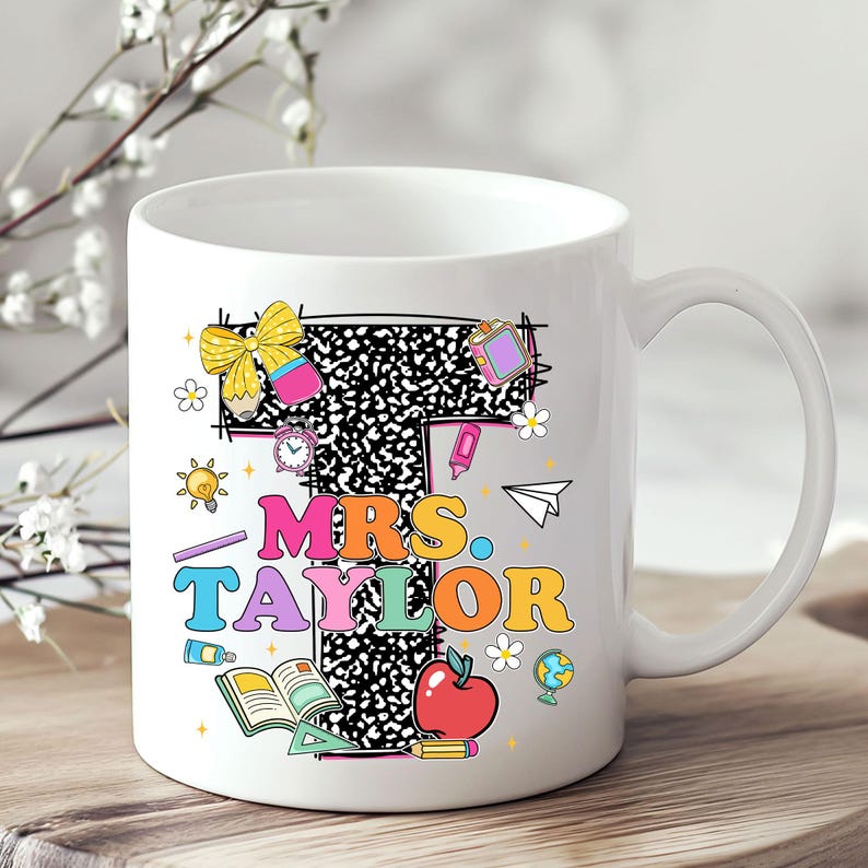 May include: White ceramic mug featuring the name "MRS. TAYLOR" in colorful, playful lettering. The design includes a large letter "T" with a black and white pattern, and various school-themed illustrations like an apple, books, and a globe.