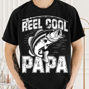 Puede incluir: Camiseta negra con un gr&aacute;fico blanco de un pez y las palabras "REEL COOL PAPA". El dise&ntilde;o incluye una ca&ntilde;a de pescar y una l&iacute;nea sobre el pez. La camiseta es de cuello redondo.