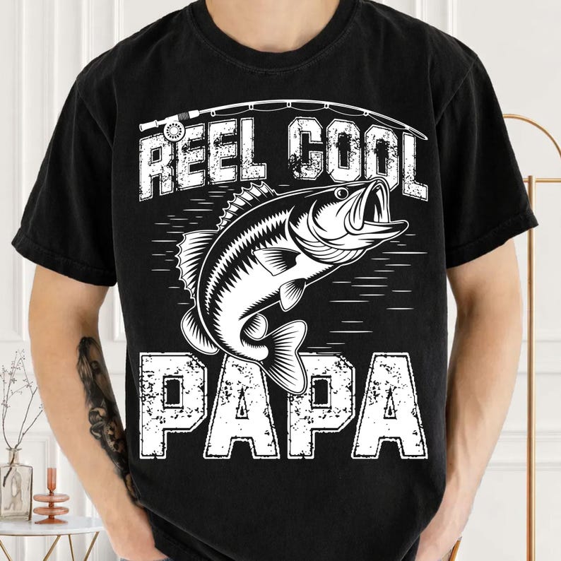 Puede incluir: Camiseta negra con un gr&aacute;fico blanco de un pez y las palabras "REEL COOL PAPA". El dise&ntilde;o incluye una ca&ntilde;a de pescar y una l&iacute;nea sobre el pez. La camiseta es de cuello redondo.