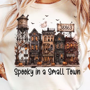 May include: A cream-colored t-shirt featuring a graphic of a spooky town with a water tower, old buildings, ghosts, bats, and pumpkins. The text "38863" appears on a billboard, and the text "Spooky in a Small Town" is printed below the illustration.
