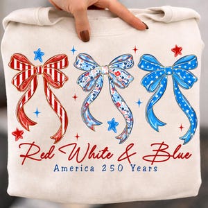 May include: Cream-colored sweatshirt featuring three decorative bows in red, white, and blue. The bows are adorned with patriotic patterns and surrounded by stars. The text "Red White & Blue America 250 Years" is printed below the bows.