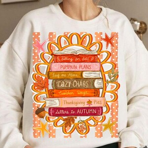 Puede incluir: Una sudadera color crema con un gr&aacute;fico de una pila de libros rodeada por un marco floral naranja. Los lomos de los libros dicen: Falling for Fall, PUMPKIN PLANS, Leaf me Alone, COZY CHAOS, Golden Days, Thanksgiving Pies y Letters to AUTUMN.