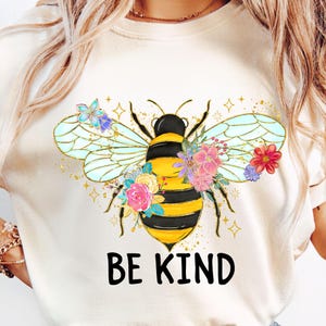 May include: Cream-colored t-shirt featuring a large, detailed bee design. The bee has yellow and black stripes, with floral accents and the words "BE KIND" in black block letters. The wings are outlined in gold.