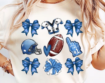 May include: A white t-shirt with blue and white football-themed graphics. The graphics include a football helmet, a football, a football jersey, a cheerleader's pom-pom, a heart, and a foam finger with the number "#1" on it.