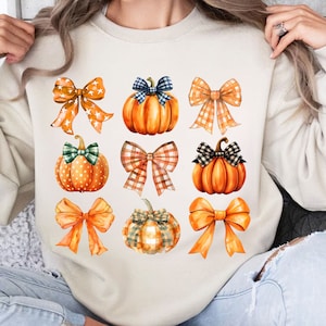 May include: A cream-colored sweatshirt with a pattern of orange pumpkins and orange, white, and brown bows. The pumpkins have green stems and some have white polka dots.