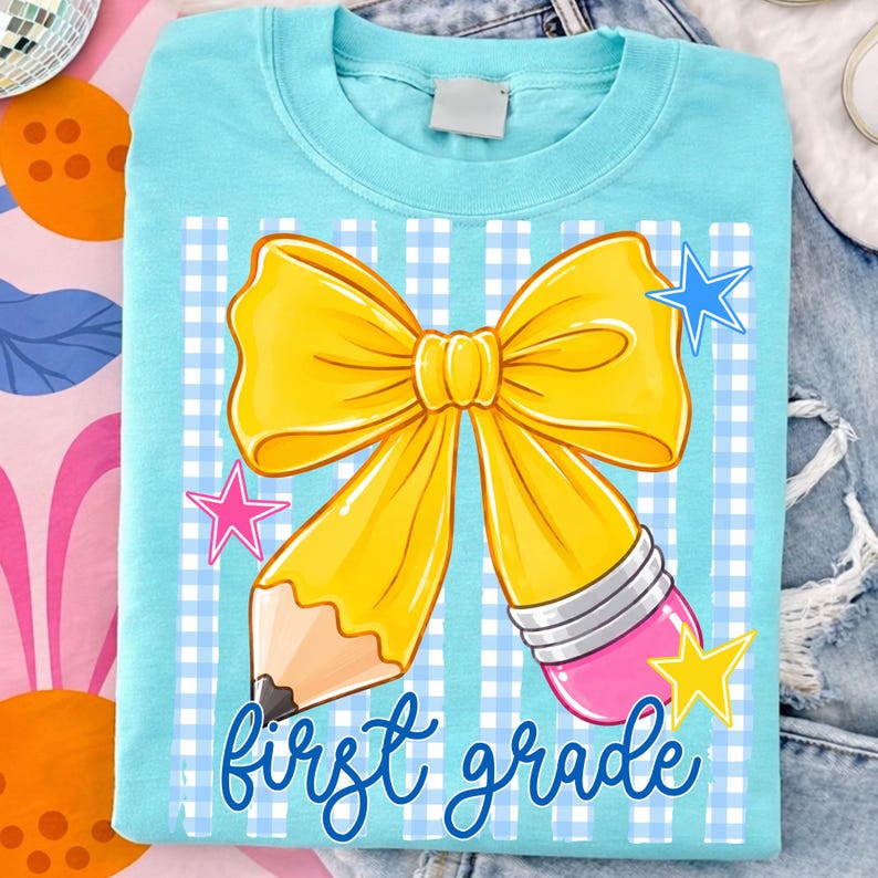 Może przedstawiać: Jasnoniebieski t-shirt z grafiką przedstawiającą dużą ż&oacute;łtą kokardę połączoną z ż&oacute;łtym oł&oacute;wkiem. Wz&oacute;r zawiera niebiesko-białe tło w kratkę vichy, małe niebieskie, r&oacute;żowe i ż&oacute;łte gwiazdki oraz napis first grade niebieską czcionką pisankową.