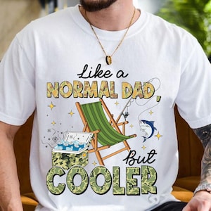 Puede incluir: Camiseta blanca con el texto "Like a NORMAL DAD But COOLER" en un diseño de camuflaje. El gráfico incluye una caña de pescar, una nevera portátil, un pez y una silla plegable verde.
