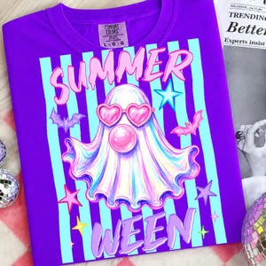 May include: A purple t-shirt with a graphic that says "SUMMER WEEN". The design features a ghost wearing heart-shaped sunglasses and a bubblegum bubble, with bats and stars. The shirt has a Comfort Colors tag.