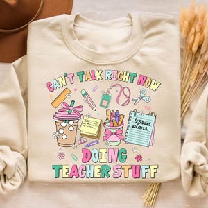 May include: Beige sweatshirt with a colorful graphic that reads "CAN'T TALK RIGHT NOW DOING TEACHER STUFF." The design includes school supplies like pencils, a ruler, scissors, and a to-go coffee cup. The text and images are in pastel colors.