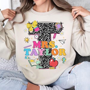 May include: Beige sweatshirt with a colorful graphic design featuring the text "MRS. TAYLOR" in various colors. The design includes school-themed elements like an apple, books, and pencils. A bow and other decorative elements complete the design.