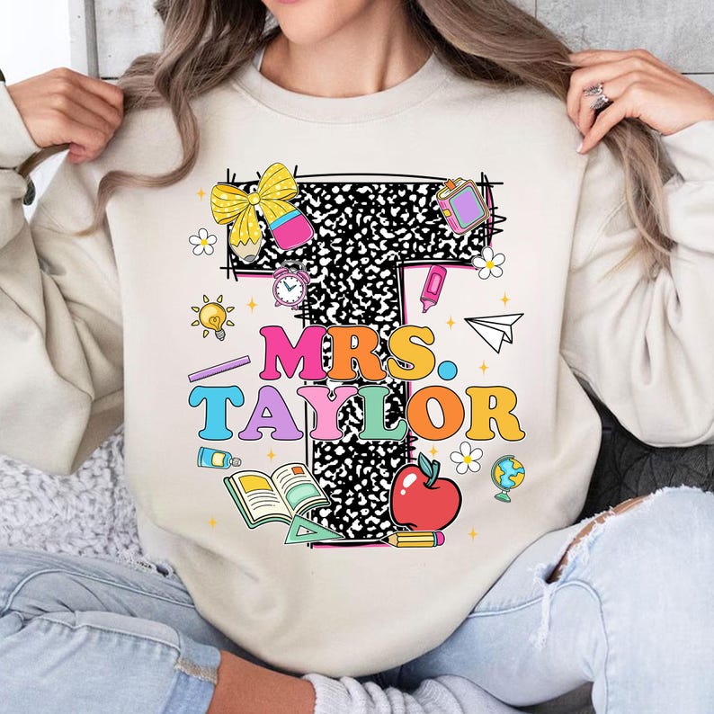 May include: Beige sweatshirt with a colorful graphic design featuring the text "MRS. TAYLOR" in various colors. The design includes school-themed elements like an apple, books, and pencils. A bow and other decorative elements complete the design.