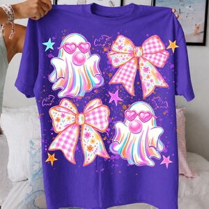 May include: A purple short-sleeve t-shirt featuring a graphic print of two ghosts wearing pink heart-shaped sunglasses and blowing bubblegum, two large decorative bows with floral and gingham patterns, small bats, and colorful stars.