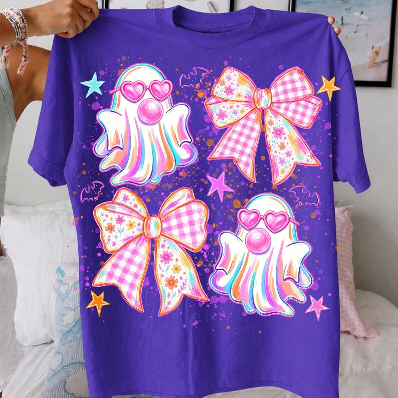 May include: A purple short-sleeve t-shirt featuring a graphic print of two ghosts wearing pink heart-shaped sunglasses and blowing bubblegum, two large decorative bows with floral and gingham patterns, small bats, and colorful stars.