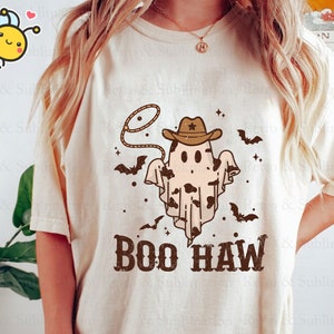 May include: A white t-shirt featuring a brown cowboy ghost wearing a brown cowboy hat and a lasso. The ghost has a black and white cow print pattern. The text "BOO HAW" is printed in brown below the ghost.