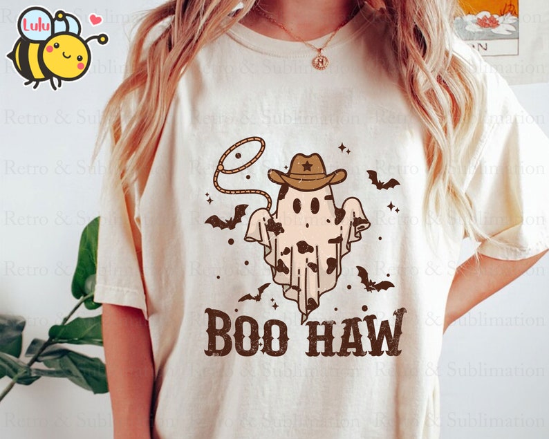 May include: A white t-shirt featuring a brown cowboy ghost wearing a brown cowboy hat and a lasso. The ghost has a black and white cow print pattern. The text "BOO HAW" is printed in brown below the ghost.