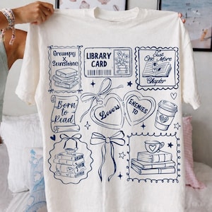 Puede incluir: Camiseta de color crema con un dise&ntilde;o gr&aacute;fico azul marino que presenta ilustraciones tem&aacute;ticas de libros y texto. El dise&ntilde;o incluye frases como "Grumpy x Sunshine", "Library Card" y "Born to Read". La camiseta es una prenda informal.