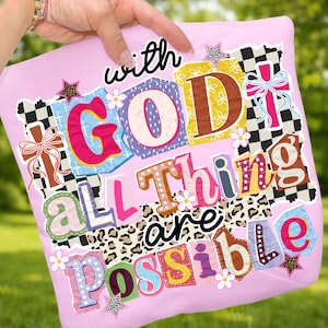 May include: A light pink t-shirt with the phrase "With God All Things Are Possible" in a colorful, patchwork design. The design includes floral and star accents.