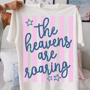 May include: Cream-colored t-shirt with a design featuring pink vertical stripes and the text "the heavens are roaring" in a blue script font. The design includes a star graphic above the text and two stars below.