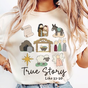 May include: A white t-shirt with a cartoon illustration of the nativity story. The illustration includes a star, a sheep, a donkey, a manger, a scroll, and three wise men. The text "True Story" and "Luke 2:1-20" is printed on the shirt.
