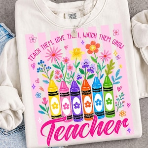 May include: Cream-colored sweatshirt with a colorful graphic. The design features a row of crayons, flowers, and the words "Teach Them, Love Them, Watch Them Grow" and "Teacher" in pink.