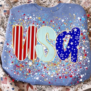 May include: A light blue sweatshirt with the word "USA" in red and white stripes, light blue, and blue with white stars. The design includes red, white, and gold paint splatters, creating a festive look.