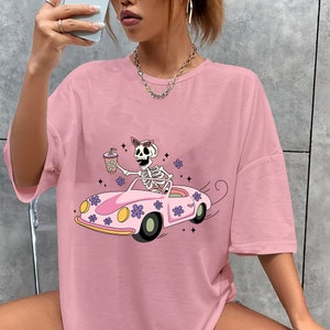 May include: Pink oversized t-shirt with a graphic of a skeleton wearing sunglasses and driving a pink car with flowers. The skeleton is holding a cup with a straw.