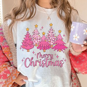 May include: White t-shirt featuring a pink and gold Christmas tree design. The trees are adorned with stars and hearts. The text "Merry Christmas" is written in pink below the trees.