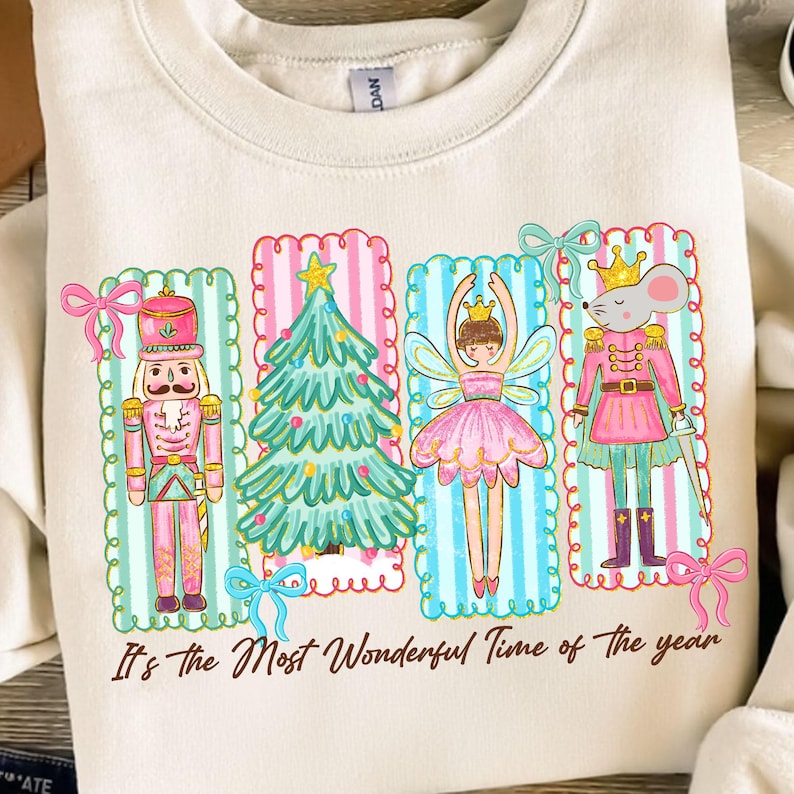 May include: Cream-colored sweatshirt featuring a festive design with a nutcracker, Christmas tree, fairy, and mouse in pink and blue frames. The phrase "It's the Most Wonderful Time of the year" is printed below the images.