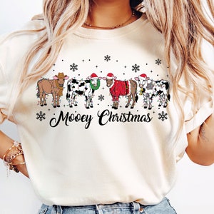 May include: A cream-coloured sweatshirt with a festive design. The graphic shows six cows decorated with Christmas lights, Santa hats, and a wreath. The phrase "Moovey Christmas" is written in a playful font below the cows. Snowflakes are scattered around the cows.