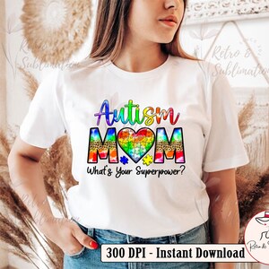 Pu&ograve; includere: Maglietta bianca con il testo "Autism Mom" in colori arcobaleno e un cuore a forma di puzzle. Il testo "What's Your Superpower?" &egrave; sotto il cuore.