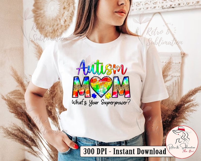 Pu&ograve; includere: Maglietta bianca con il testo "Autism Mom" in colori arcobaleno e un cuore a forma di puzzle. Il testo "What's Your Superpower?" &egrave; sotto il cuore.