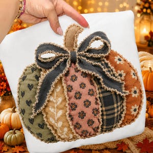 May include: A white throw pillow featuring a central fabric pumpkin applique. The pumpkin is composed of five vertical segments in olive green with hearts, tan with leaves, pink with flowers, black plaid, and orange with daisies, topped with a dark bow.