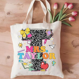 May include: Cream-colored tote bag with the name "MRS. TAYLOR" in colorful, retro-style lettering. The design includes school-themed illustrations like an apple, books, and a globe. The bag has long, cream-colored handles and is decorated with a bouquet of pink tulips.