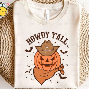 May include: A white t-shirt with the text "Howdy Y'all" in brown lettering. A cartoon pumpkin wearing a cowboy hat is in the centre of the shirt. The pumpkin is orange with a brown hat and a brown bandana.