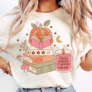 May include: A white t-shirt with a cute illustration of a pumpkin reading a book. The pumpkin is wearing glasses and has a stack of books next to it. There is a candle next to the pumpkin with the text "Fallen Leaves & Apple Cider".