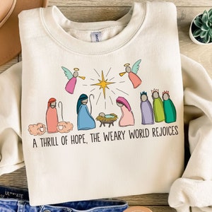 Puede incluir: Sudadera de color crema con una colorida ilustración de la escena de la Natividad, con ángeles, la estrella y los tres reyes magos. El texto "A THRILL OF HOPE, THE WEARY WORLD REJOICES" está impreso debajo. La sudadera es de material suave.