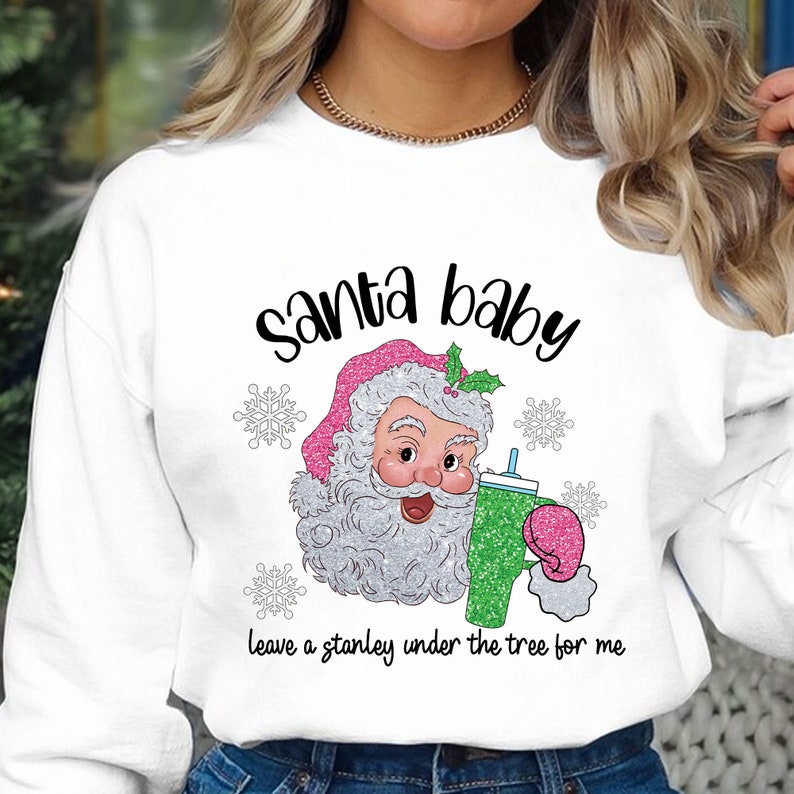May include: White sweatshirt featuring a graphic of Santa Claus holding a green drink with a pink lid. The text "Santa baby leave a stanley under the tree for me" is printed below the graphic.
